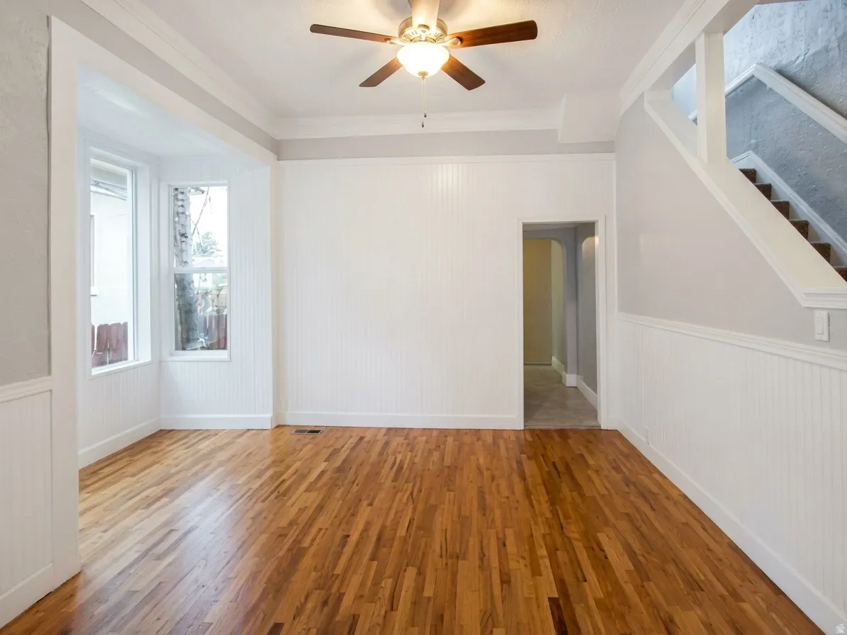 Hardwood flooring throughout a room featuring a bay window, beadboard wainscoting, and a ceiling fan with light fixture