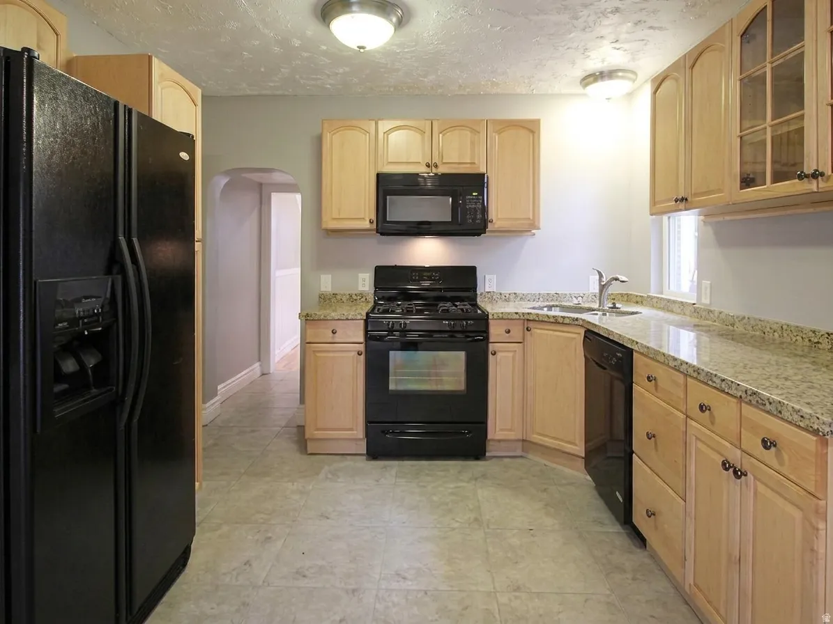 Kitchen featuring light wood cabinetry, stone countertops, black appliances, a double basin sink, and tile flooring