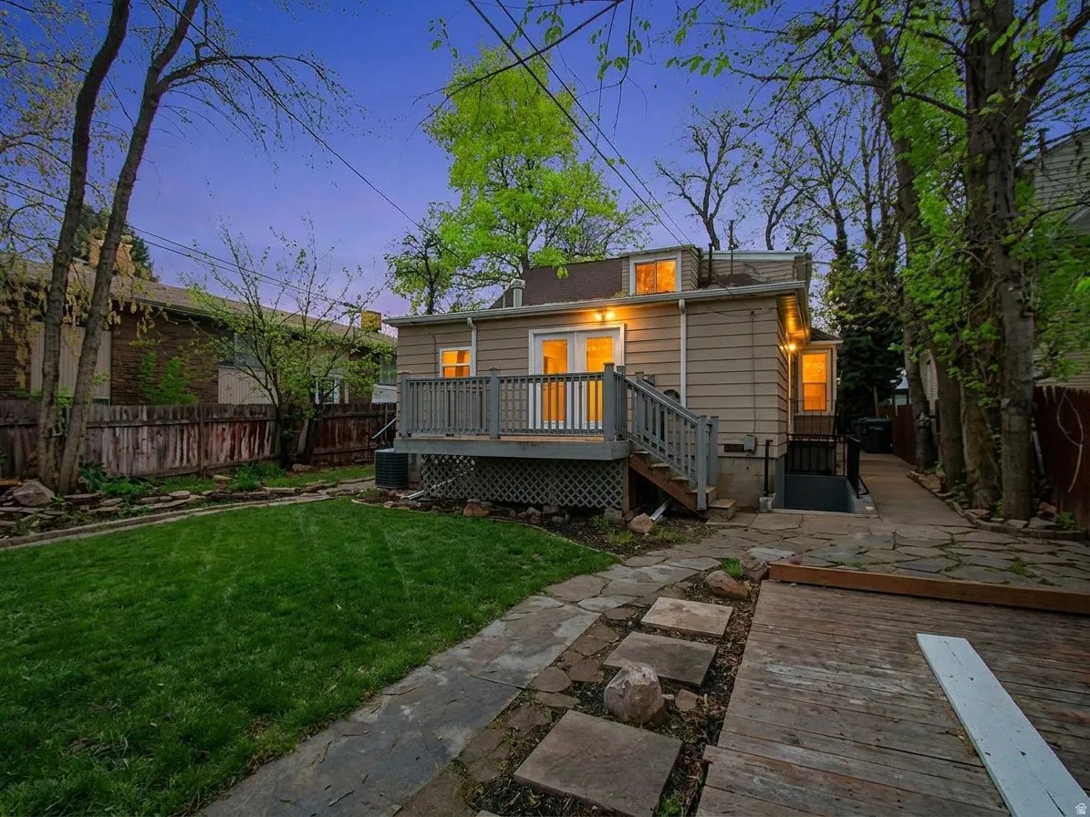 Rear exterior featuring a wood deck with railing and stairs, double glass doors, a stone paver pathway, and a grassy area