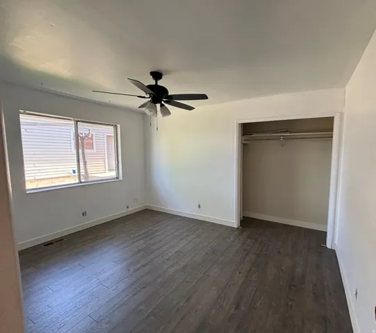Spacious room featuring wood-finish flooring, a ceiling fan with integrated lighting, a large window, and an open closet with shelving and hanging rods