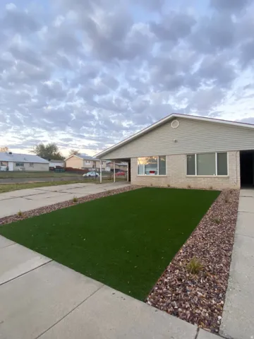 Front exterior featuring a gabled roofline, light-toned brick facade, siding accents, artificial turf lawn, and decorative gravel landscaping