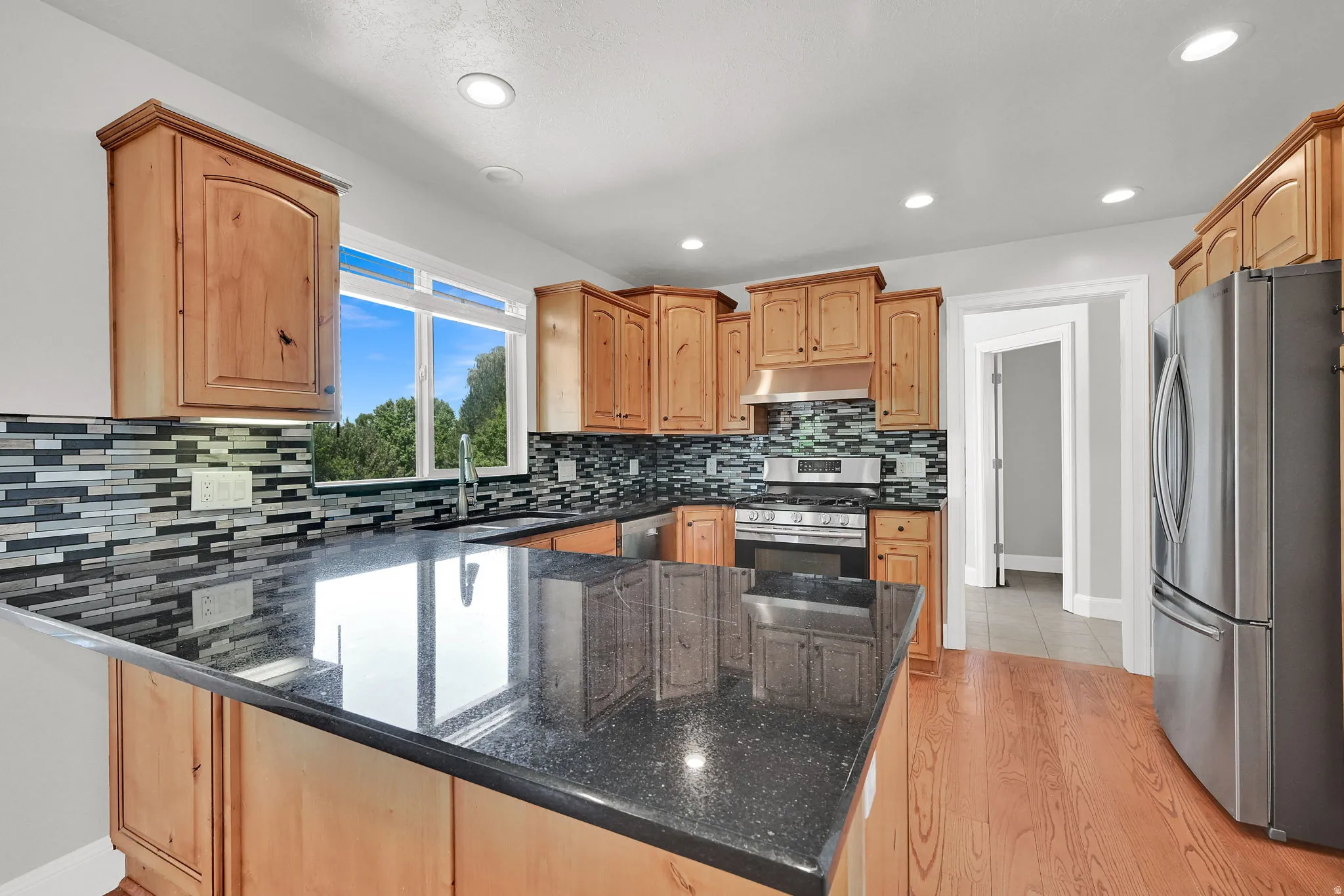 Kitchen featuring a central island with a black speckled countertop, natural wood cabinetry, stainless steel appliances, mosaic tile backsplash, and wood-finish flooring