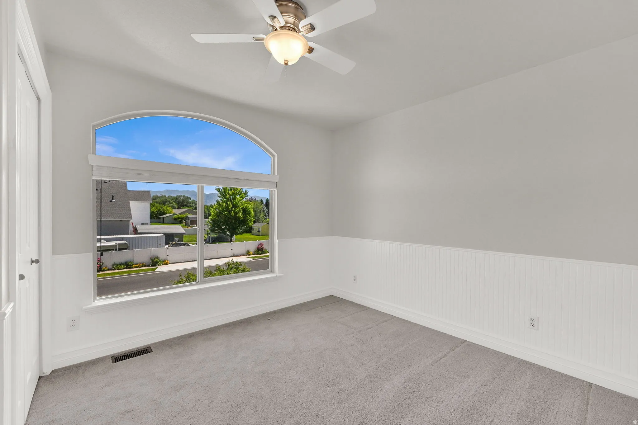 Arched window with broad sills, white beadboard wainscoting, light gray walls, ceiling fan with integrated lighting, and gray carpeting