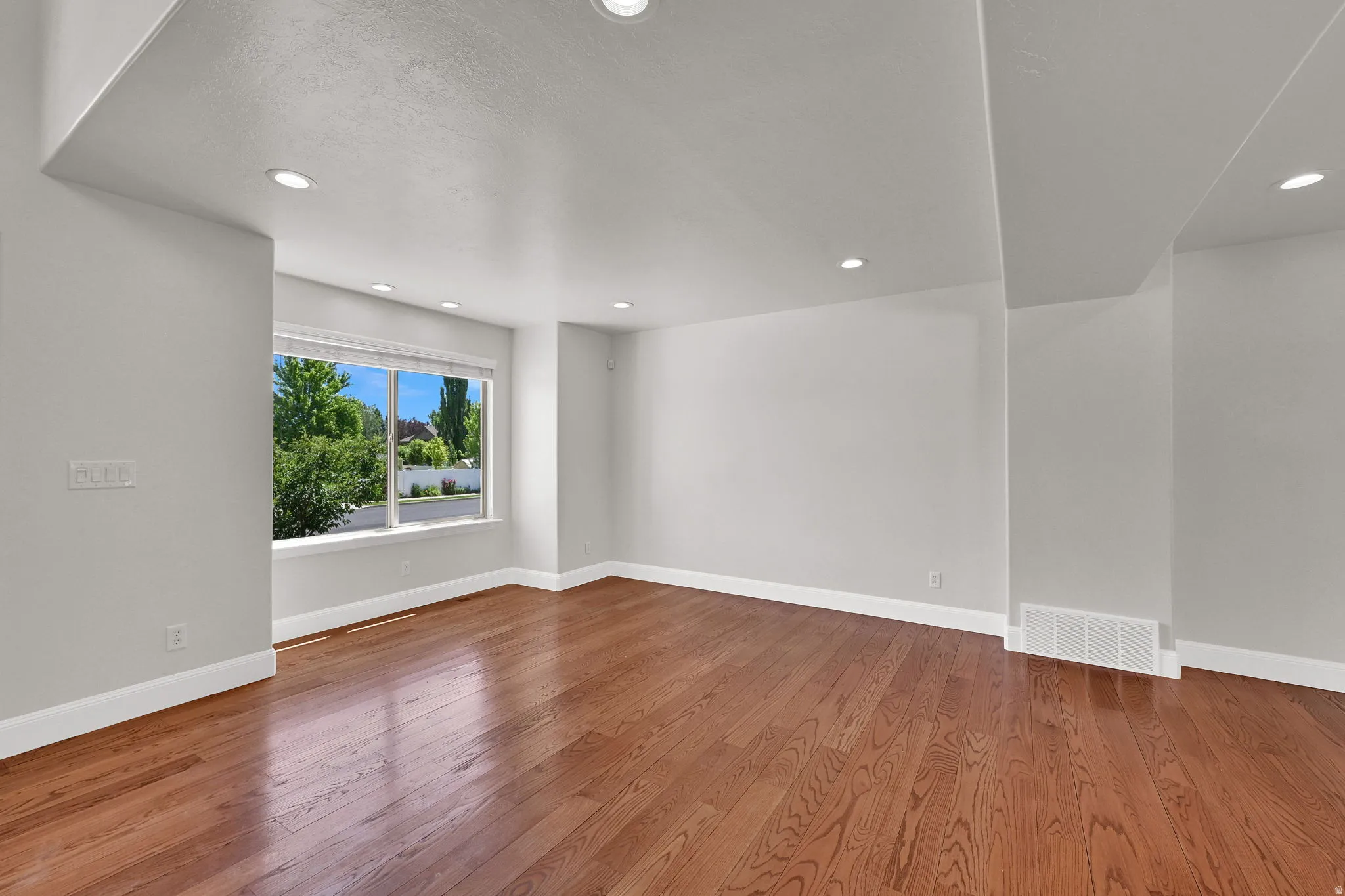 Spacious room featuring rich wood-finish flooring, recessed ceiling lighting, a large window with white trim, white baseboards, and light gray wall paint