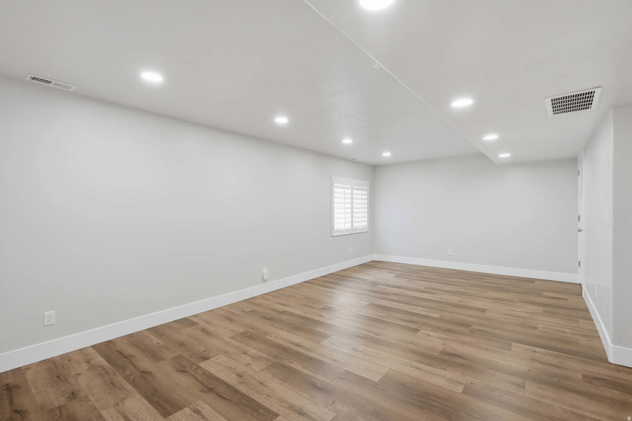 Spacious interior featuring wood-finish flooring, recessed lighting, a window with white plantation shutters, white baseboards, and a white ceiling
