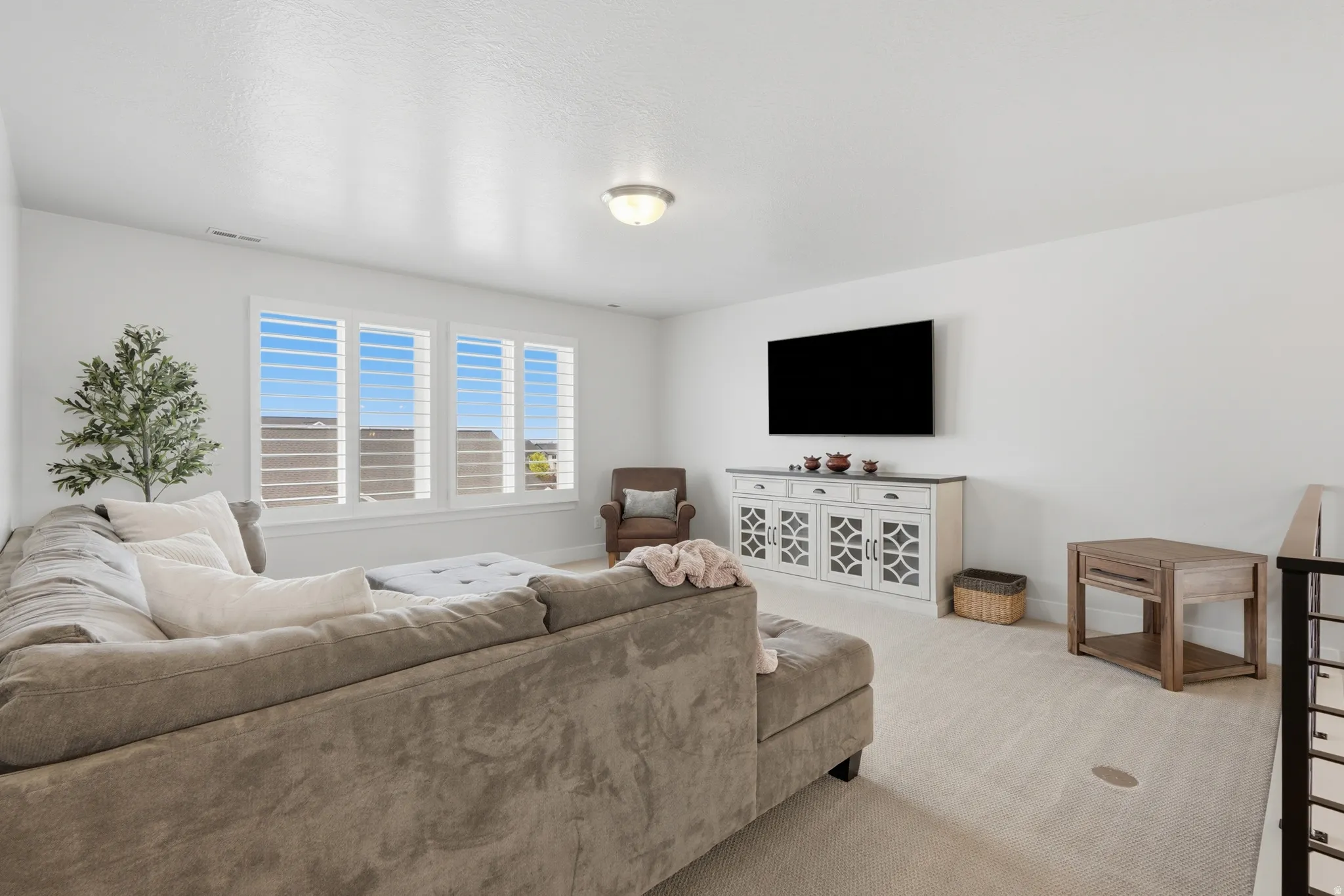 Spacious room featuring multiple windows with louvered shutters, light-toned carpeting, a wall-mounted television, and a built-in console cabinet with decorative glass panels