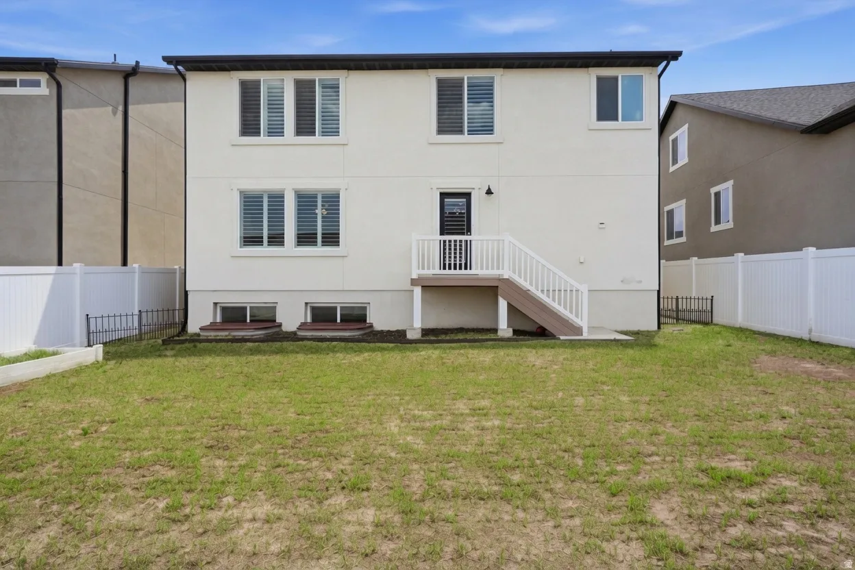 Rear exterior featuring a two-story design with light-colored stucco, multiple windows with white frames, a black rear door, and a white-railed deck with stairs descending to a green lawn