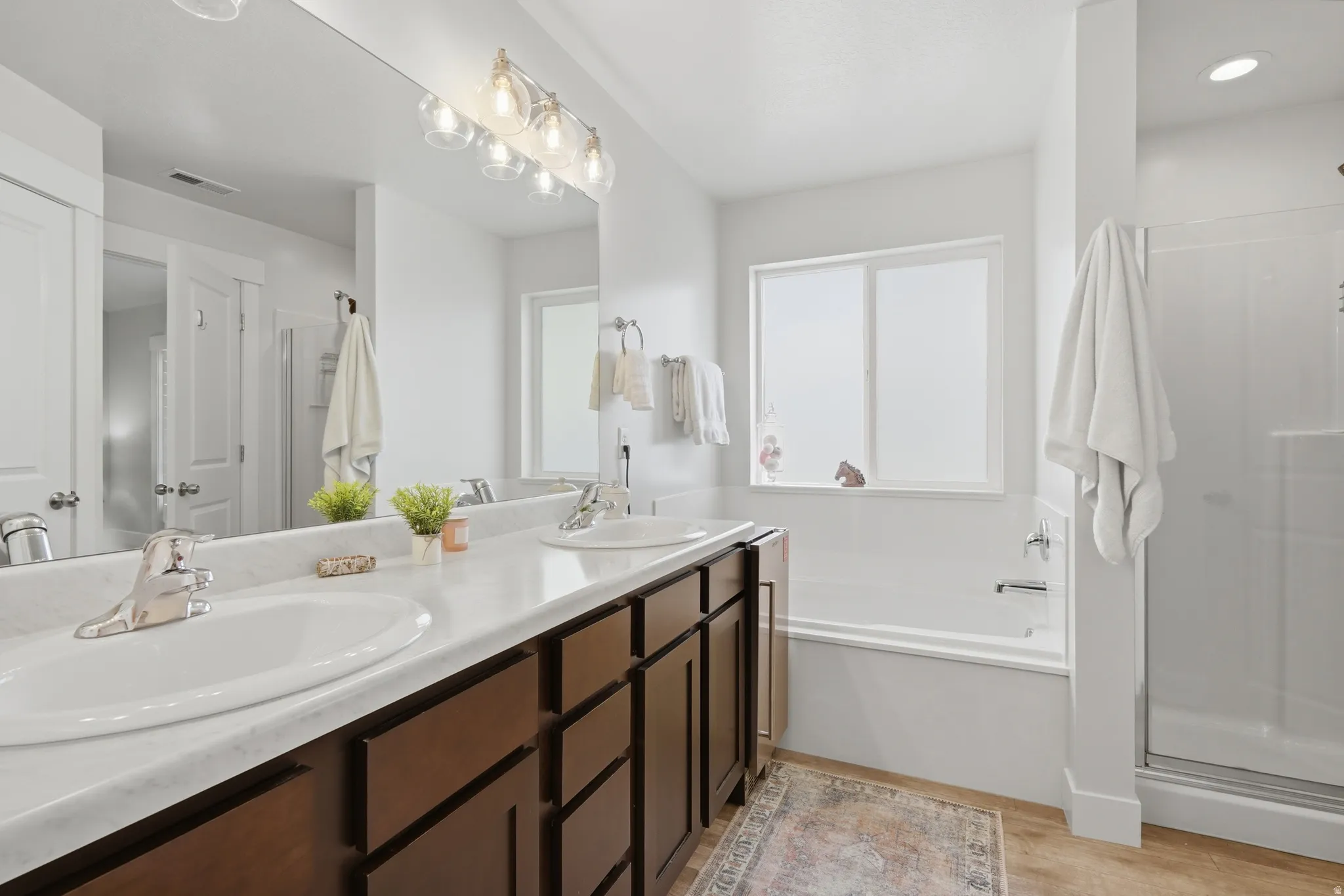 Bathroom featuring a dual-sink vanity with white countertops, a large mirror, a light fixture with clear glass shades, a built-in bathtub, and a separate shower enclosure