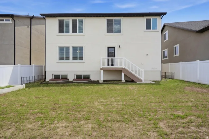Rear exterior featuring a two-story design with light-colored stucco, multiple windows with white frames, a black rear door, and a white-railed deck with stairs descending to a green lawn
