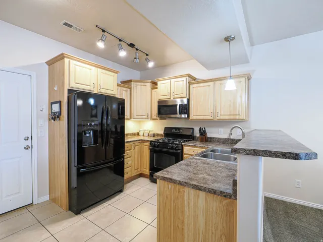 Kitchen featuring natural wood-finish cabinetry, black appliance suite, track lighting, a dual basin stainless steel sink, and a breakfast bar with a stone-finish countertop