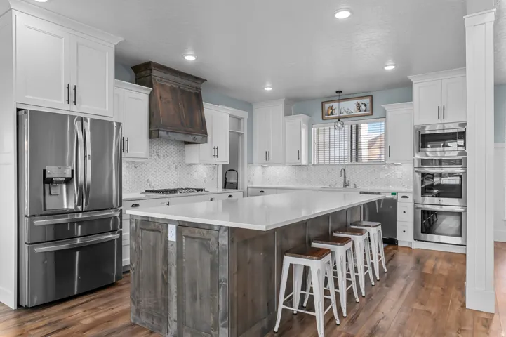 Expansive kitchen featuring a large central island with a light-toned countertop, white shaker-style cabinetry, stainless steel appliances, a tile backsplash, and wood-finish flooring