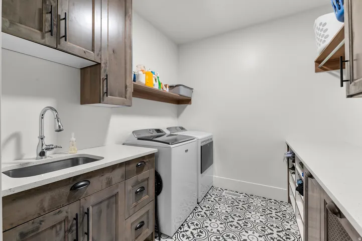 Laundry area featuring patterned floor tile, wood-finish cabinetry with matte black hardware, white countertops, and a stainless steel sink with a chrome faucet