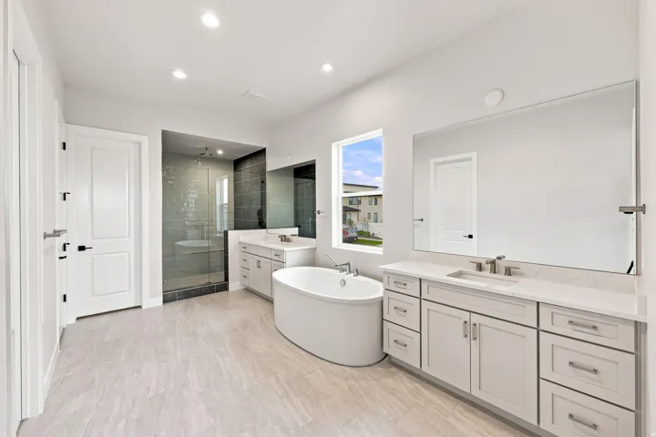 Freestanding soaking tub, dual vanities with white countertops, large glass-enclosed shower, and light-toned tile flooring