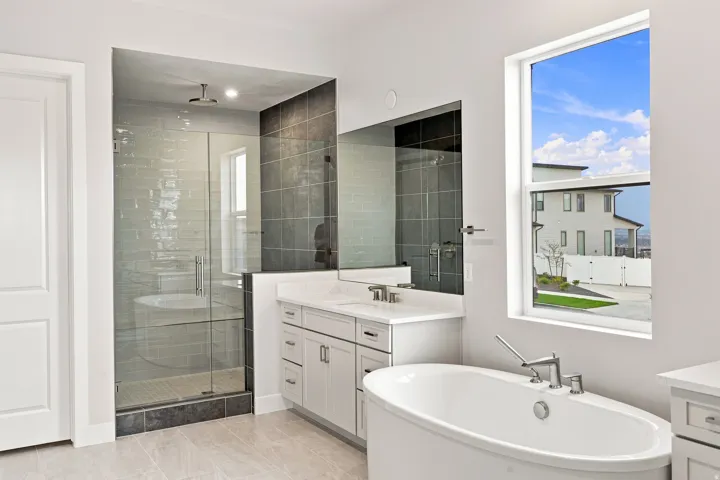 Freestanding oval soaking tub, glass-enclosed shower with tiled walls, dual vanity with undermount sinks, and a large window