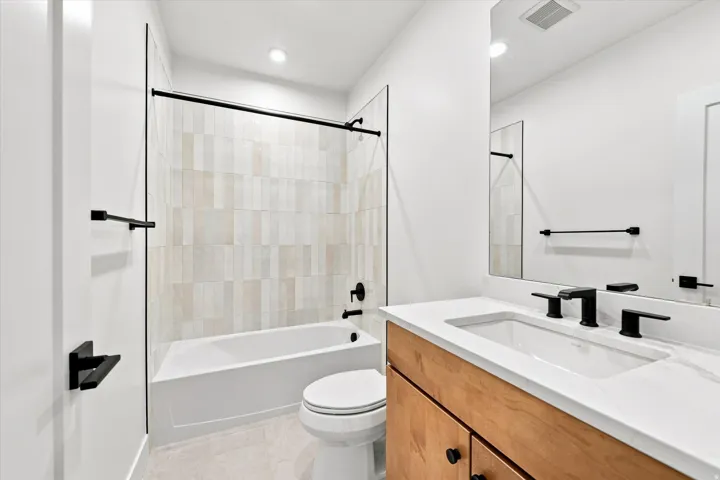 White rectangular bathtub with matte black fixtures, vertical tile surround, and a wood-finish vanity with an integrated sink and large mirror