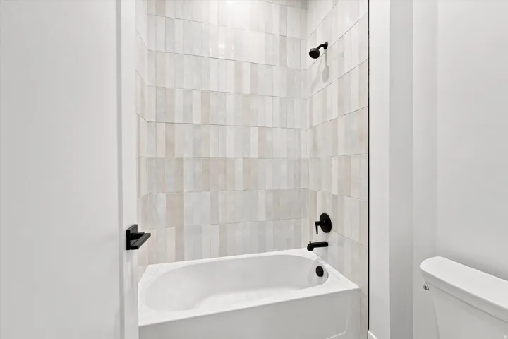Recessed bathtub with vertically oriented tile surround, matte black shower head, matte black tub filler, and a toilet