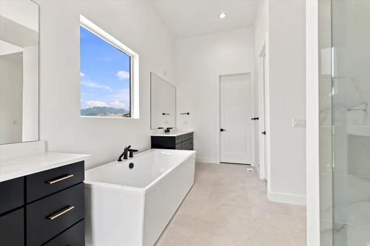 Freestanding soaking tub with matte black fixtures, dual vanities with dark cabinetry and light countertops, large format tile flooring, and a glass-enclosed shower