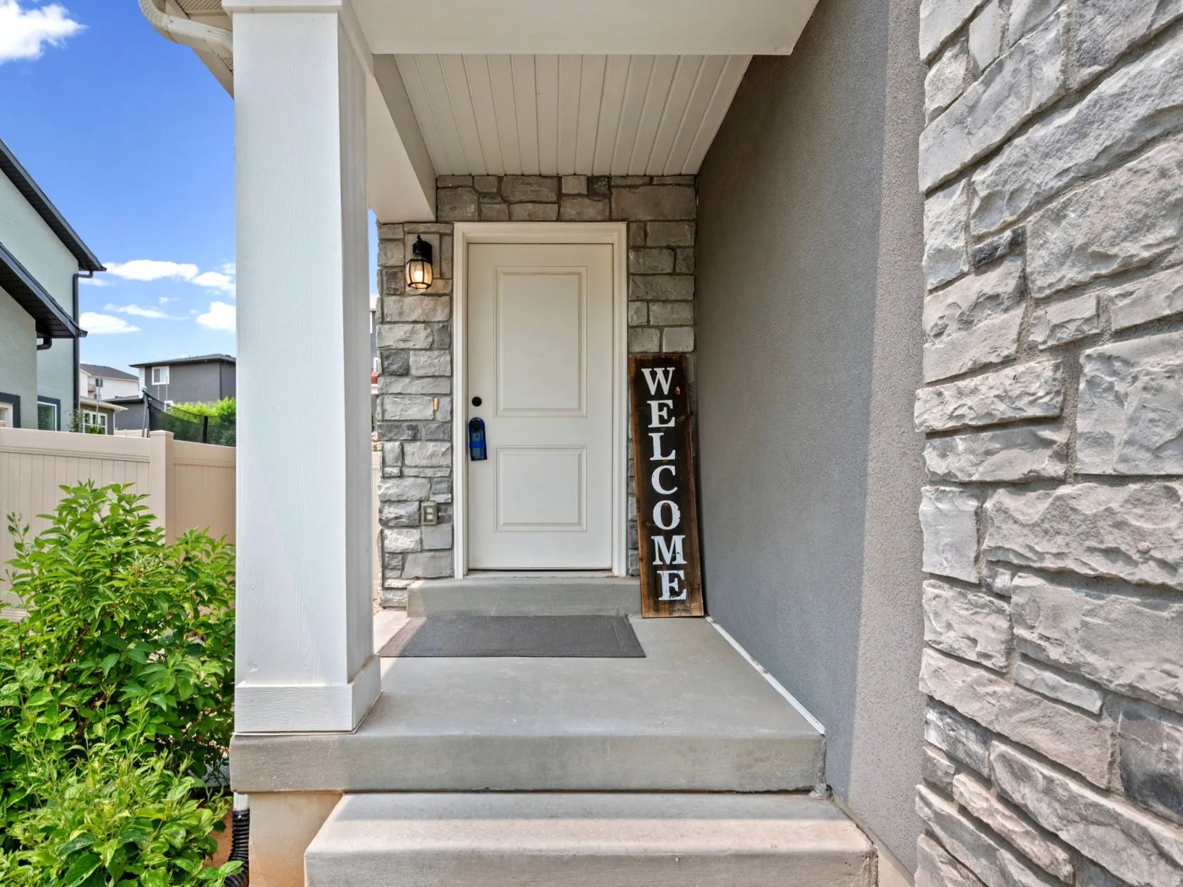 Entryway featuring a white paneled door, stone accent siding, concrete steps, a covered porch with a white column, and exterior lighting