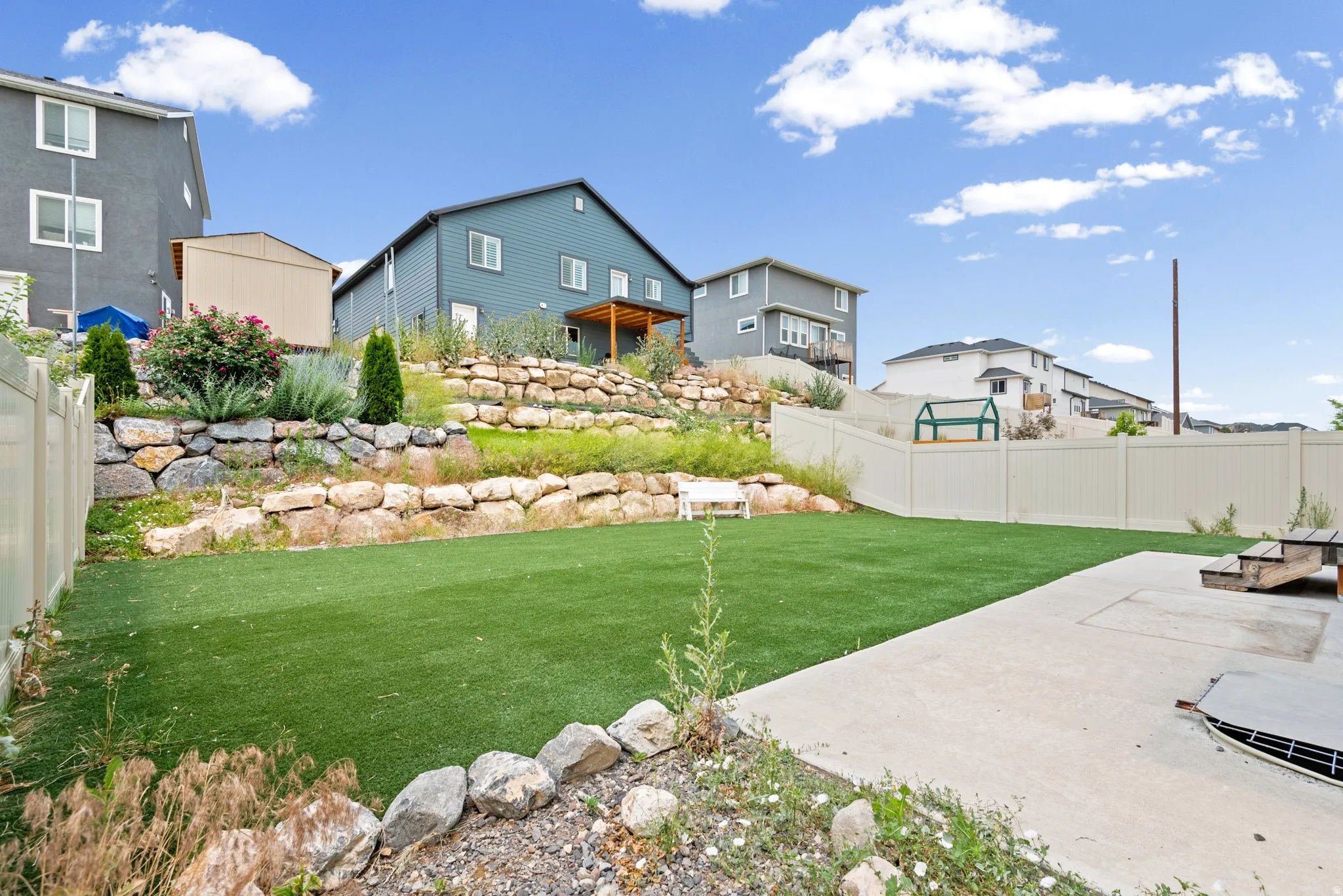 Expansive backyard featuring artificial turf, a concrete patio, and tiered rock retaining walls with established landscaping