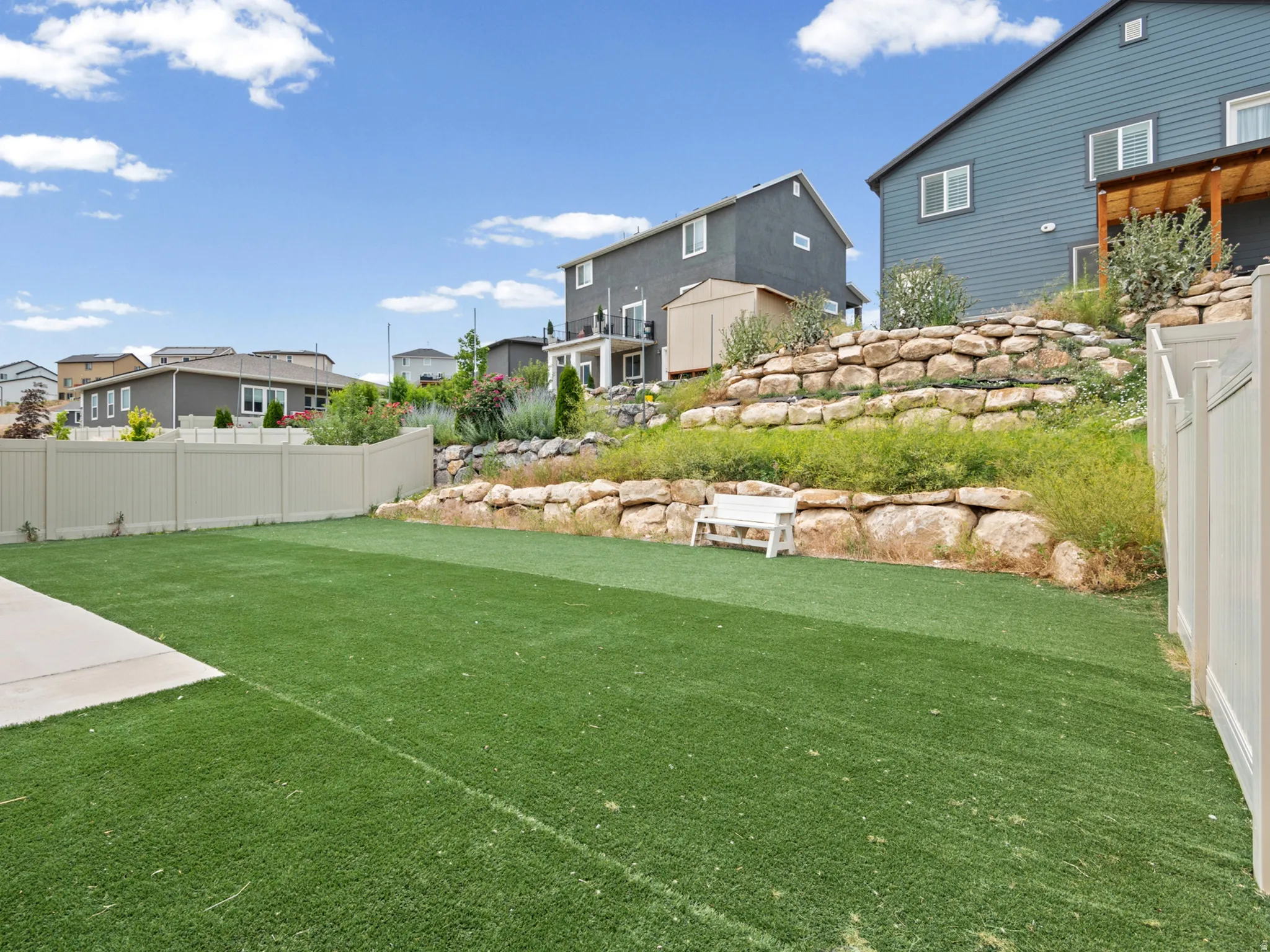 Spacious backyard featuring artificial turf, a concrete patio, and a tiered rock retaining wall