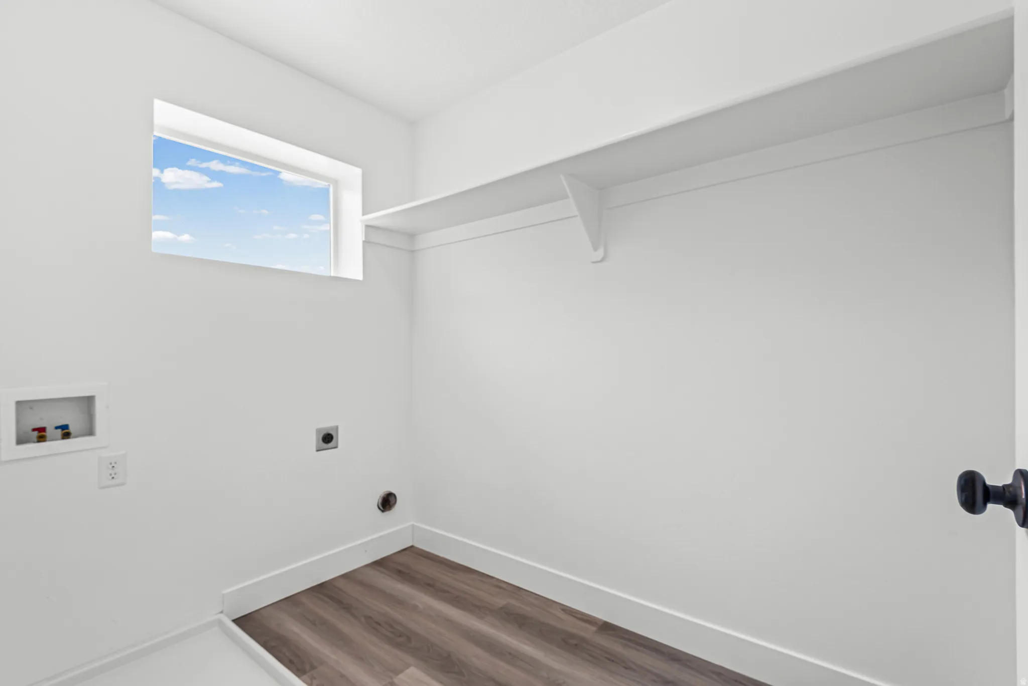 Dedicated utility space featuring bright white walls, wood-finish flooring, a high window, an integrated wall shelf, and a black door handle