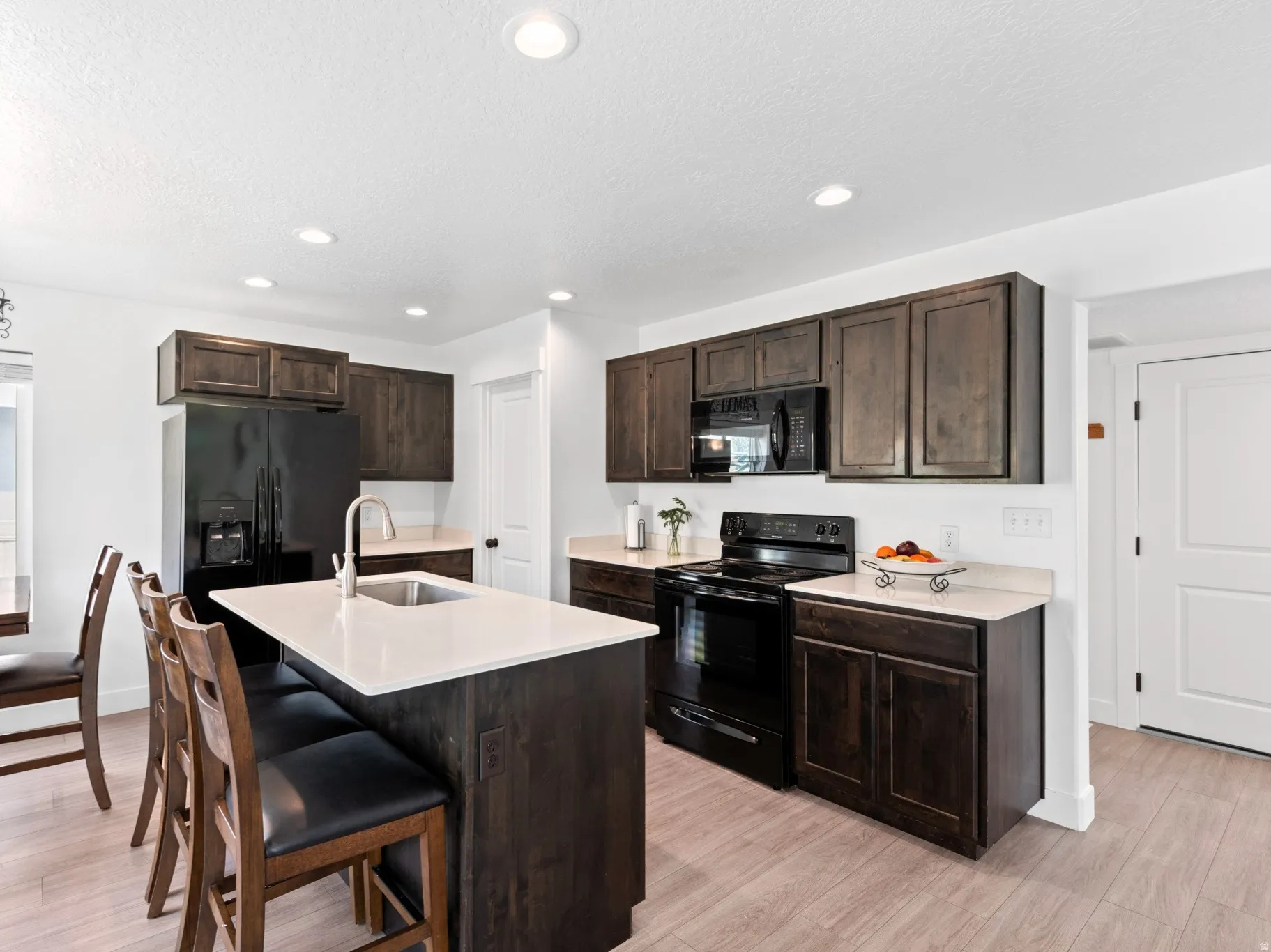 Kitchen featuring an island with a stainless steel sink and light-toned countertop, dark wood-finish cabinetry, black appliances including a refrigerator, range, and built-in microwave, and light wood-finish flooring
