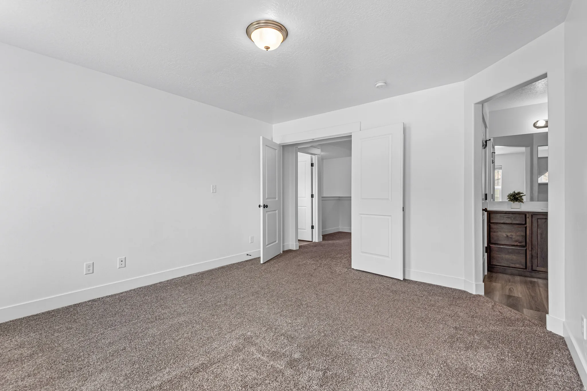 Carpeted room with white walls, a semi-flush mount ceiling light, and an entry to an additional space with wood-finish flooring and a dark wood vanity with a light-colored countertop