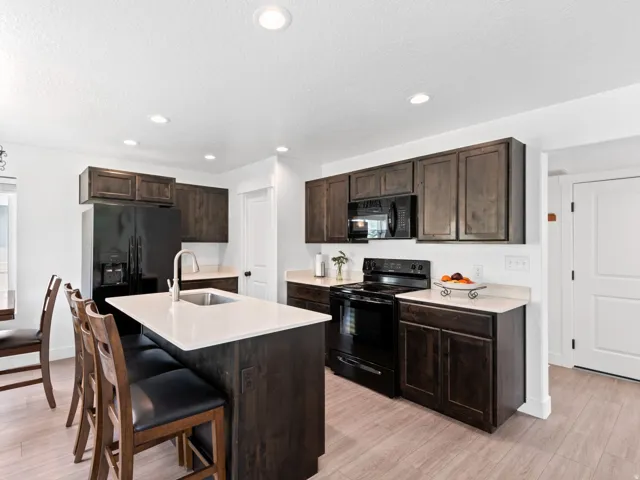Kitchen featuring an island with a stainless steel sink and light-toned countertop, dark wood-finish cabinetry, black appliances including a refrigerator, range, and built-in microwave, and light wood-finish flooring