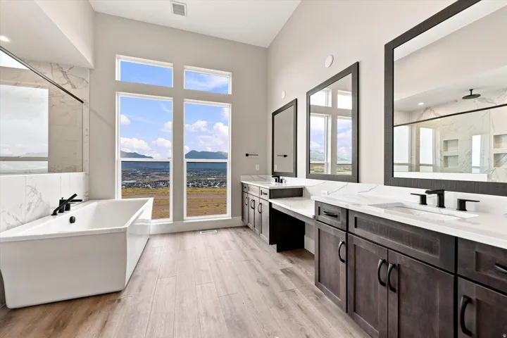 Freestanding soaking tub with black fixtures, expansive windows, dual vanity with undermount sinks, wood-finish flooring, and a glass-enclosed shower