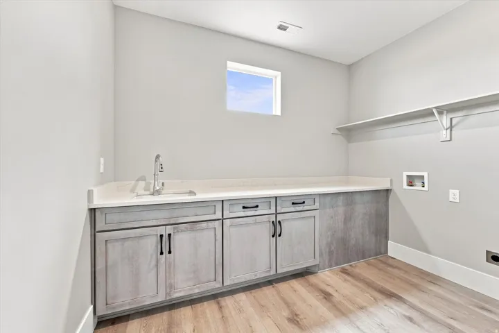 Light wood-finish flooring, built-in shelving, white countertop, undermount sink with a gooseneck faucet, and light gray cabinetry with dark hardware