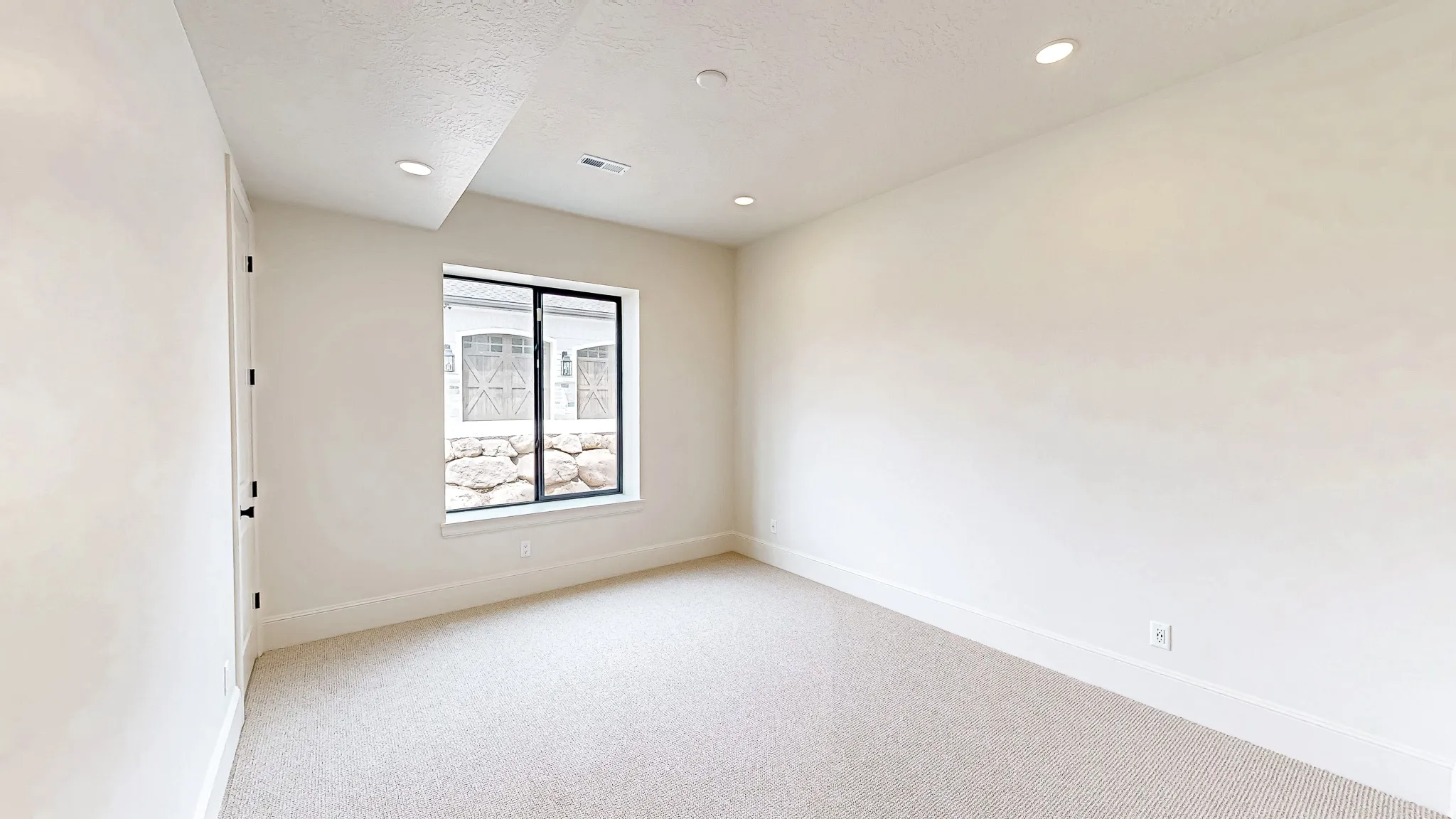 Recessed lighting illuminates this room featuring light-colored carpeting, white walls, and a window with black trim