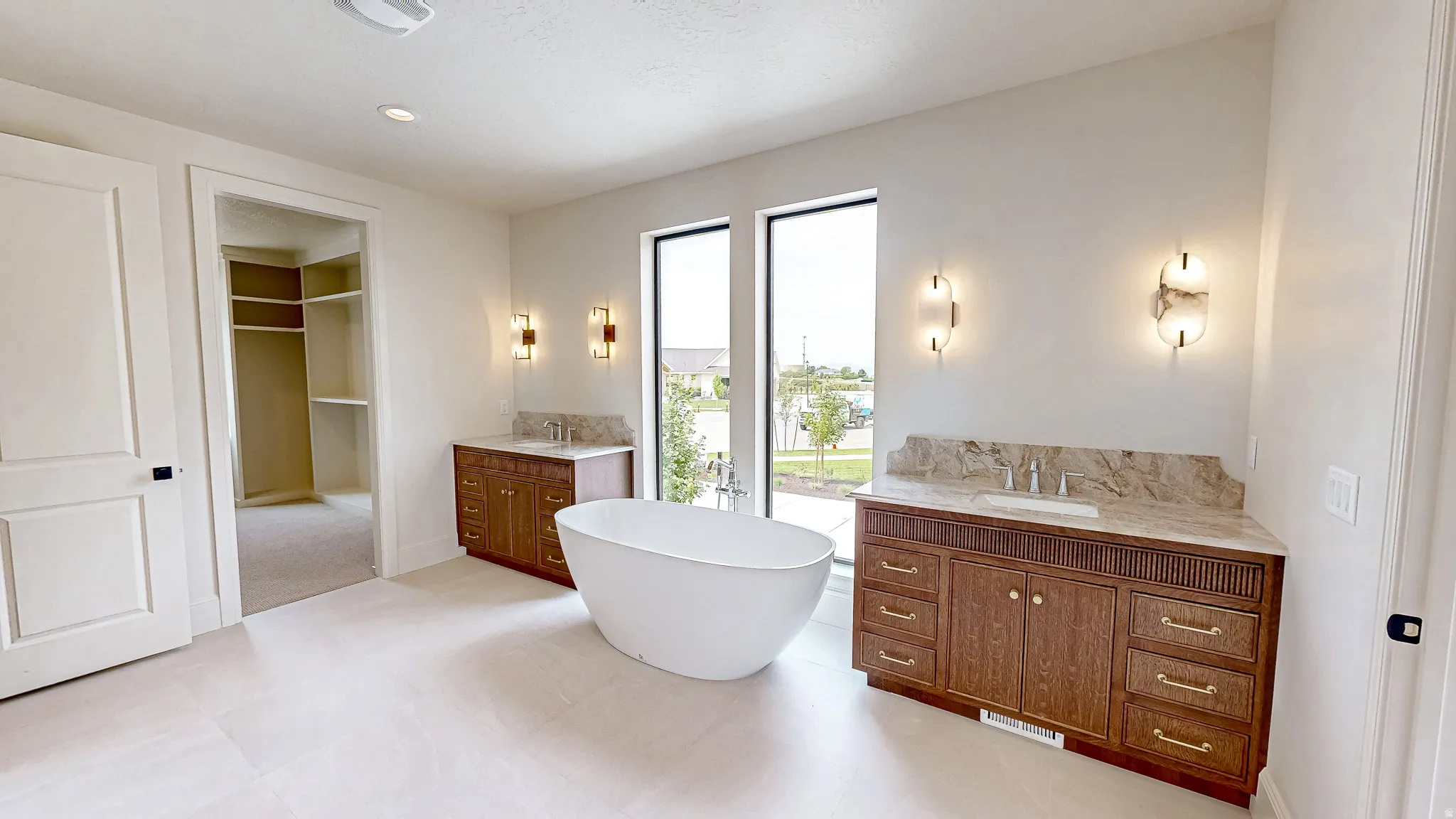 Spacious bathroom featuring a freestanding soaking tub, dual vanity cabinets with stone countertops, modern wall sconces, and large windows