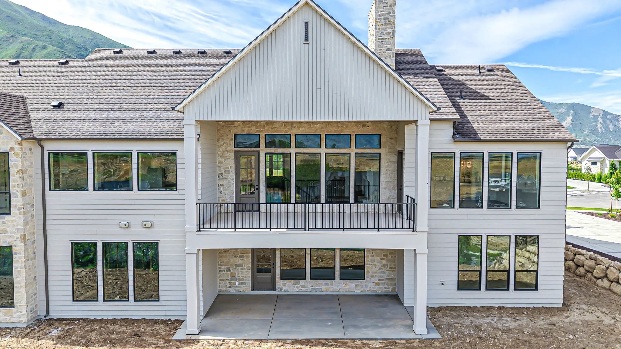 Multi-level exterior featuring light-toned siding, stone accents, multiple large windows, a covered balcony with a black railing, and a ground-level covered patio
