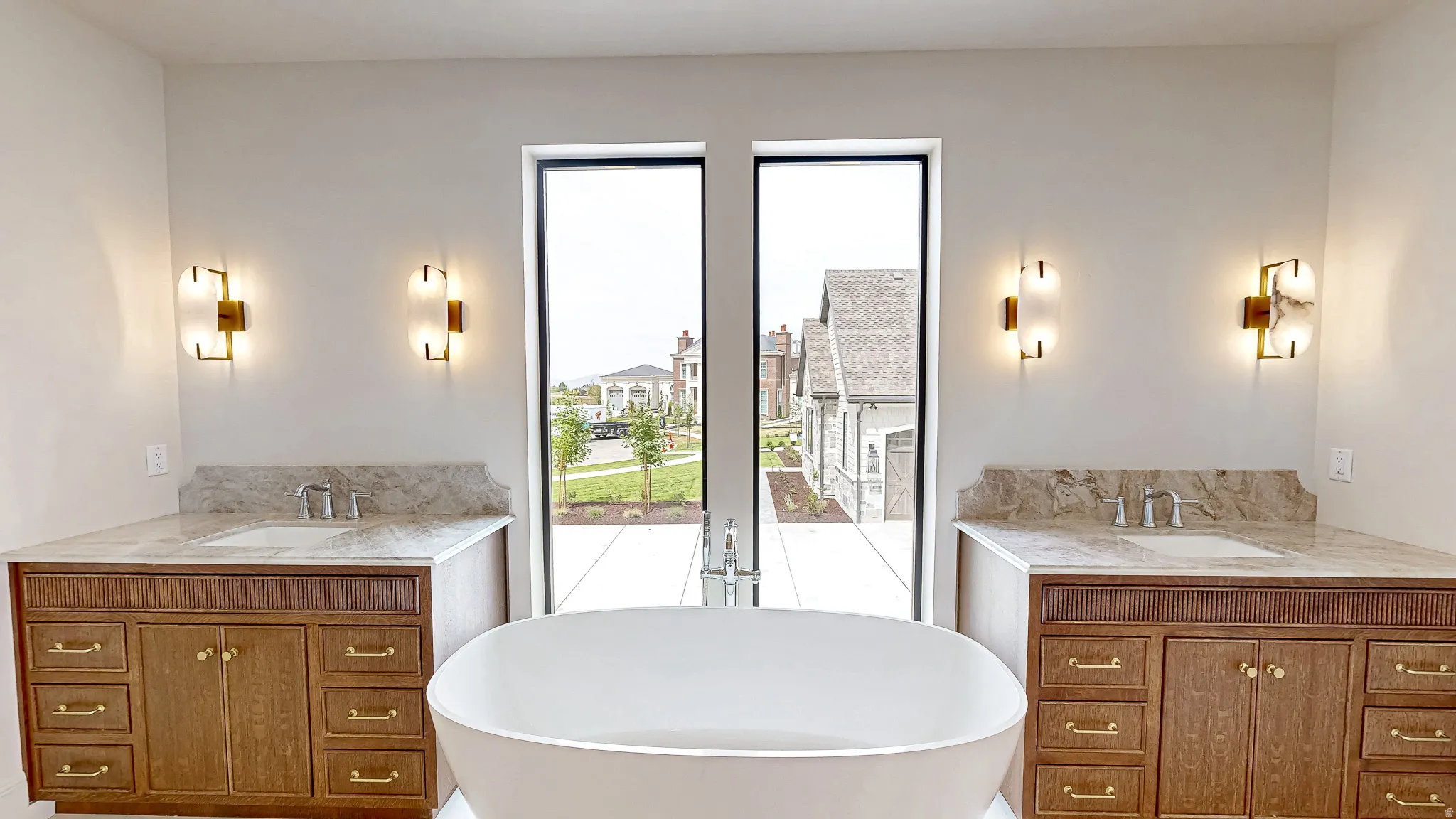 Bathroom featuring a freestanding white soaking tub, dual vanity cabinets with gold-tone hardware, stone countertops with integrated sinks, and wall-mounted sconce lighting