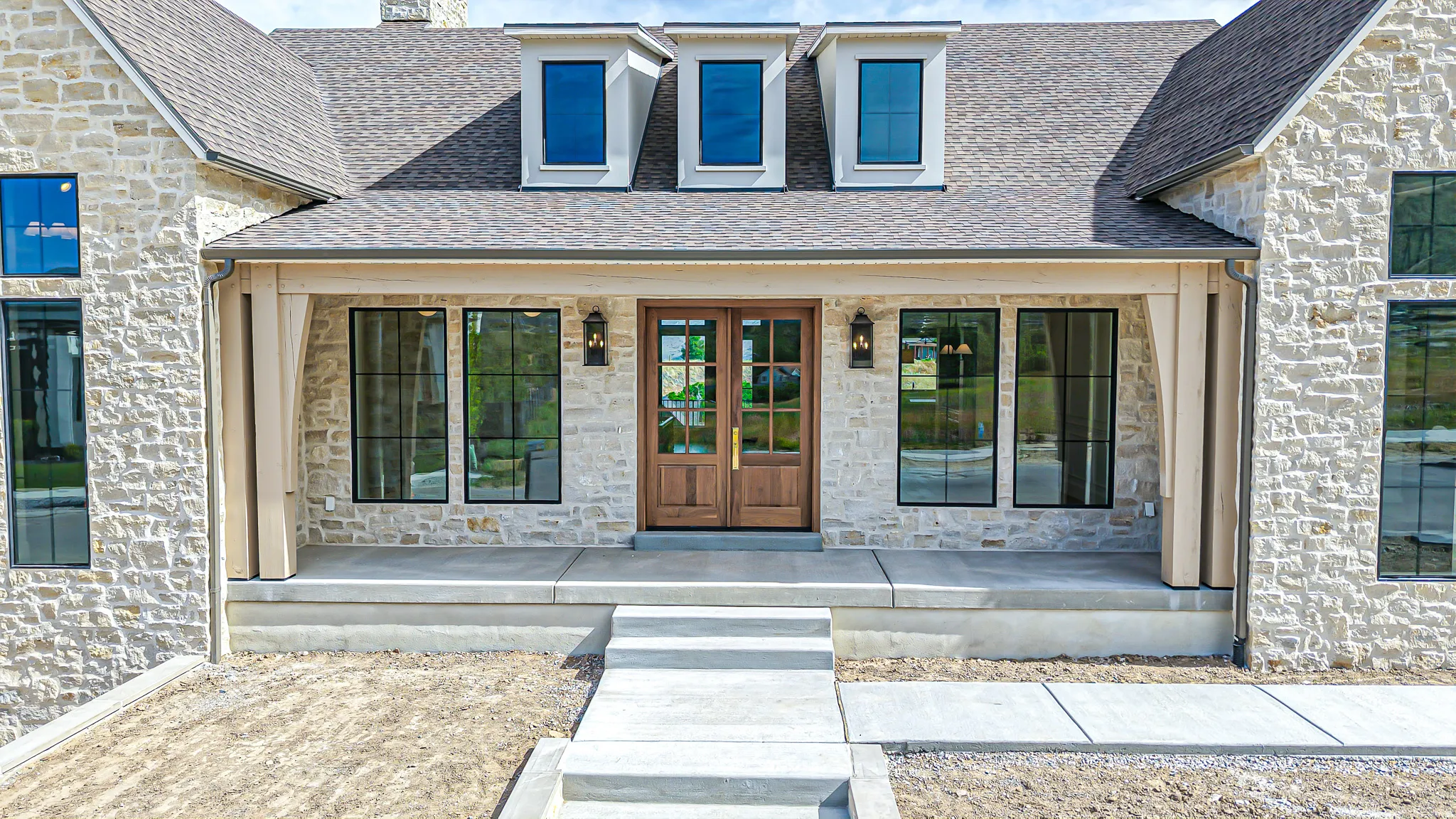 Stone facade home featuring a prominent front entry with double wood-finish doors, a covered porch with exposed wood beams, black-framed windows, and multiple dormer windows