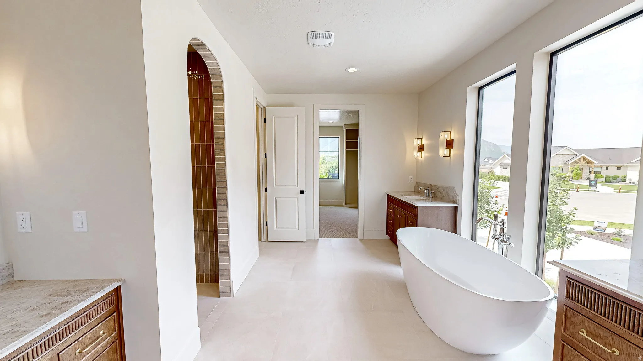 Spacious bathroom featuring a freestanding soaking tub, large windows, an arched shower entry with vertical tile, a double vanity with stone countertop, and wood-finish cabinetry