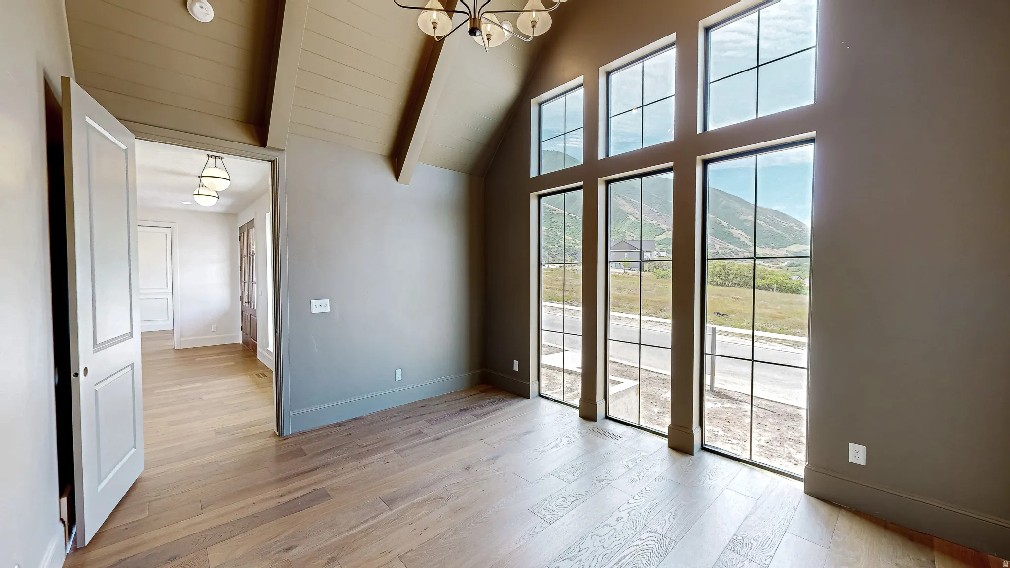 Room featuring multiple floor-to-ceiling windows with black grids, wood-finish flooring, a vaulted ceiling with exposed beams, and a prominent chandelier