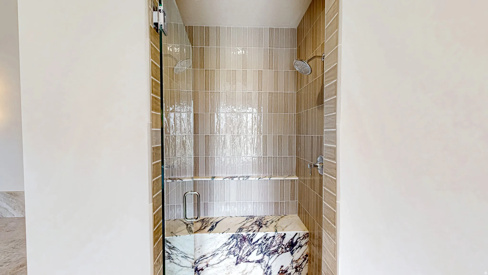Walk-in shower featuring vertical beige and white tile, two showerheads, a built-in bench with marble-like veining, and a glass enclosure