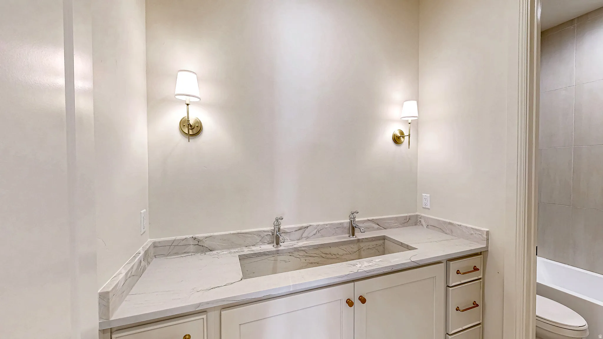 Dual vanity with an integrated basin, white cabinetry, brass hardware, and wall-mounted sconces