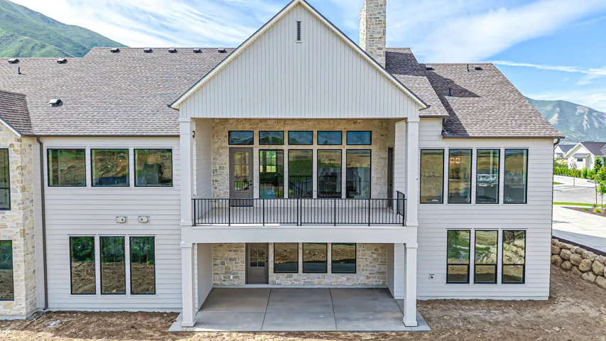Multi-level exterior featuring light-toned siding, stone accents, multiple large windows, a covered balcony with a black railing, and a ground-level covered patio