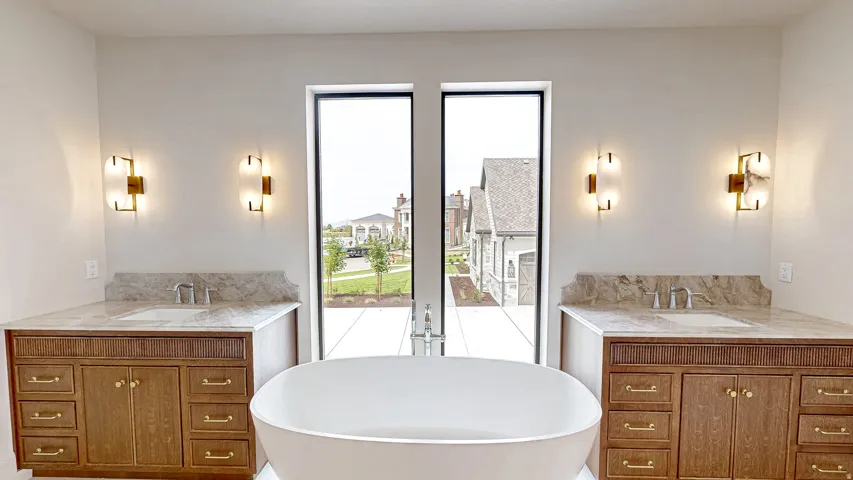 Bathroom featuring a freestanding white soaking tub, dual vanity cabinets with gold-tone hardware, stone countertops with integrated sinks, and wall-mounted sconce lighting