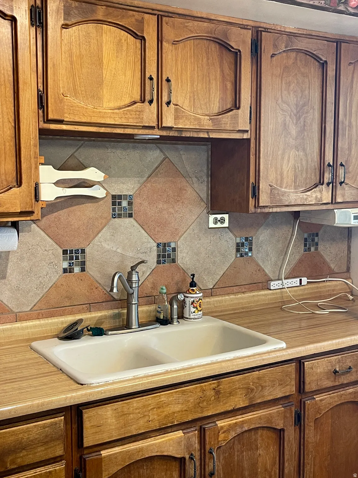 Kitchen sink area featuring light wood-finish cabinetry, a neutral-toned countertop, and a tiled backsplash with decorative accents
