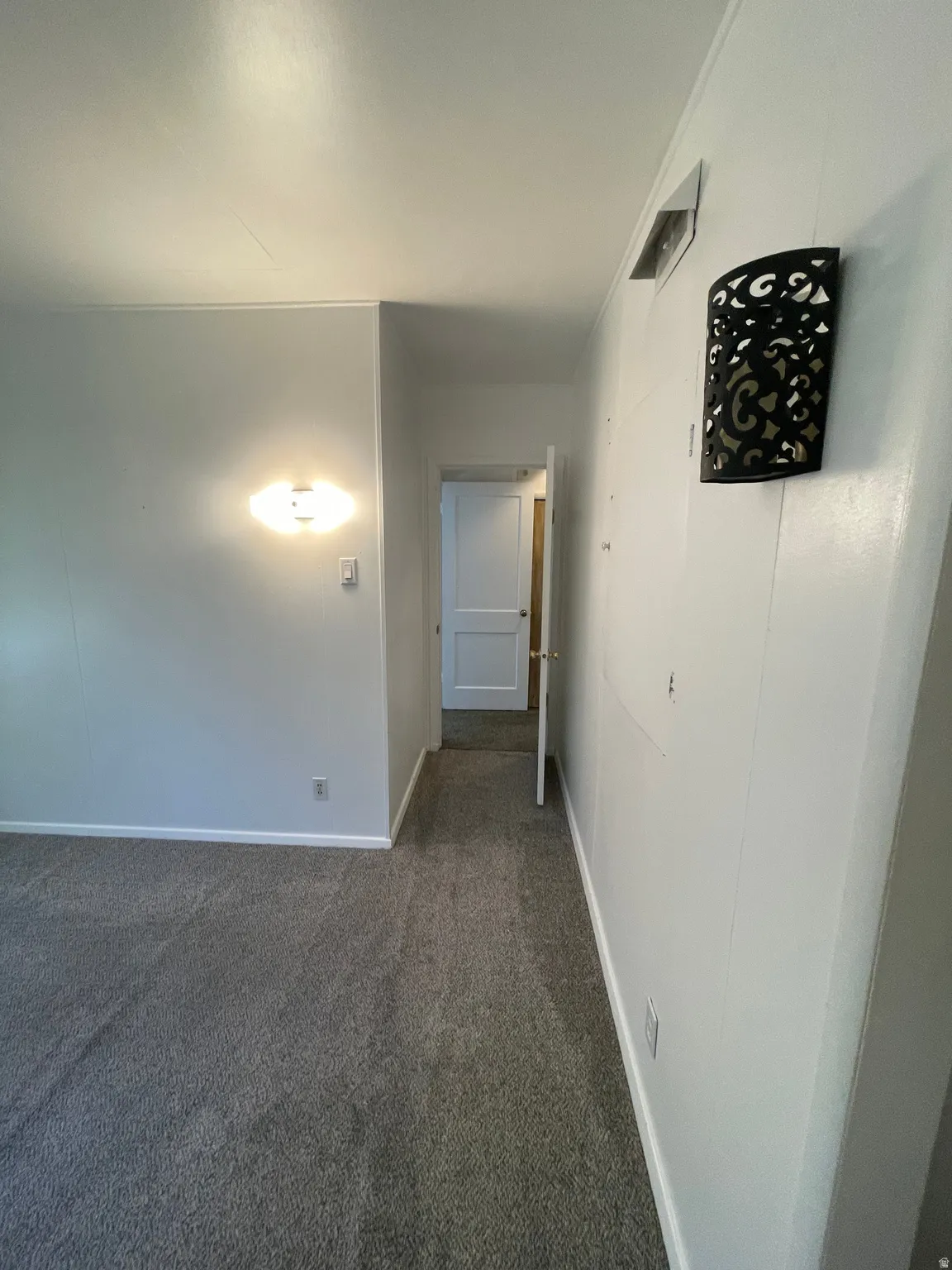 Hallway featuring gray carpet flooring, white walls, a white door, and a decorative black wall sconce