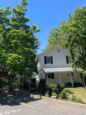 White exterior featuring black shutters, a covered porch with a railing, and surrounding mature trees