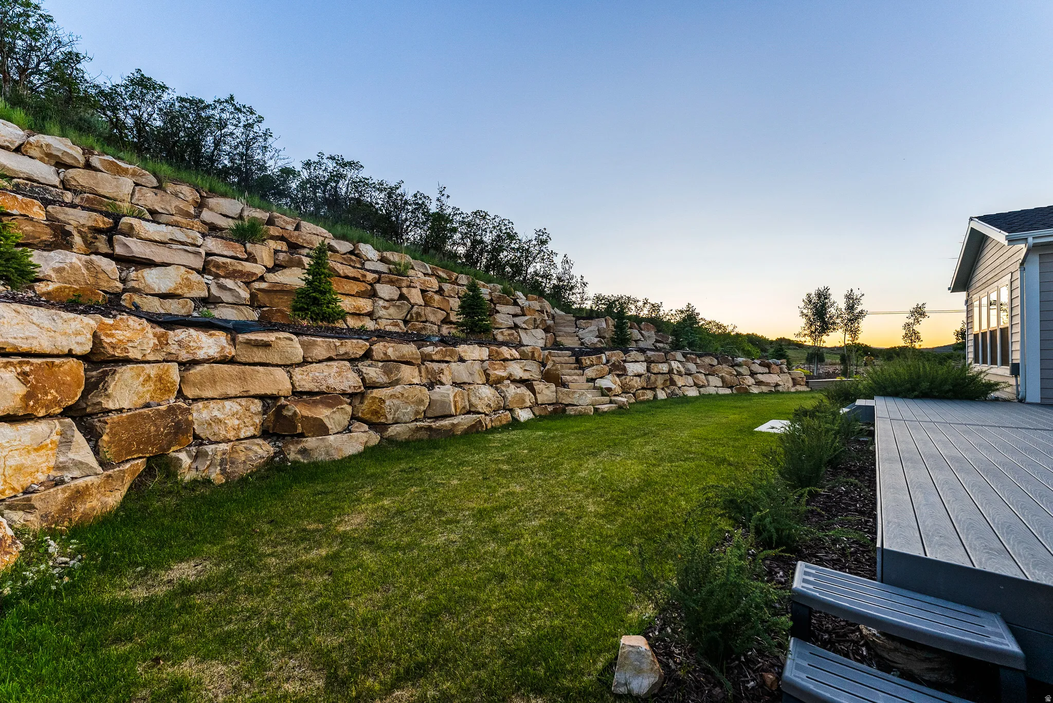 Outdoor area featuring a stone retaining wall, green lawn, composite decking, and mature landscaping