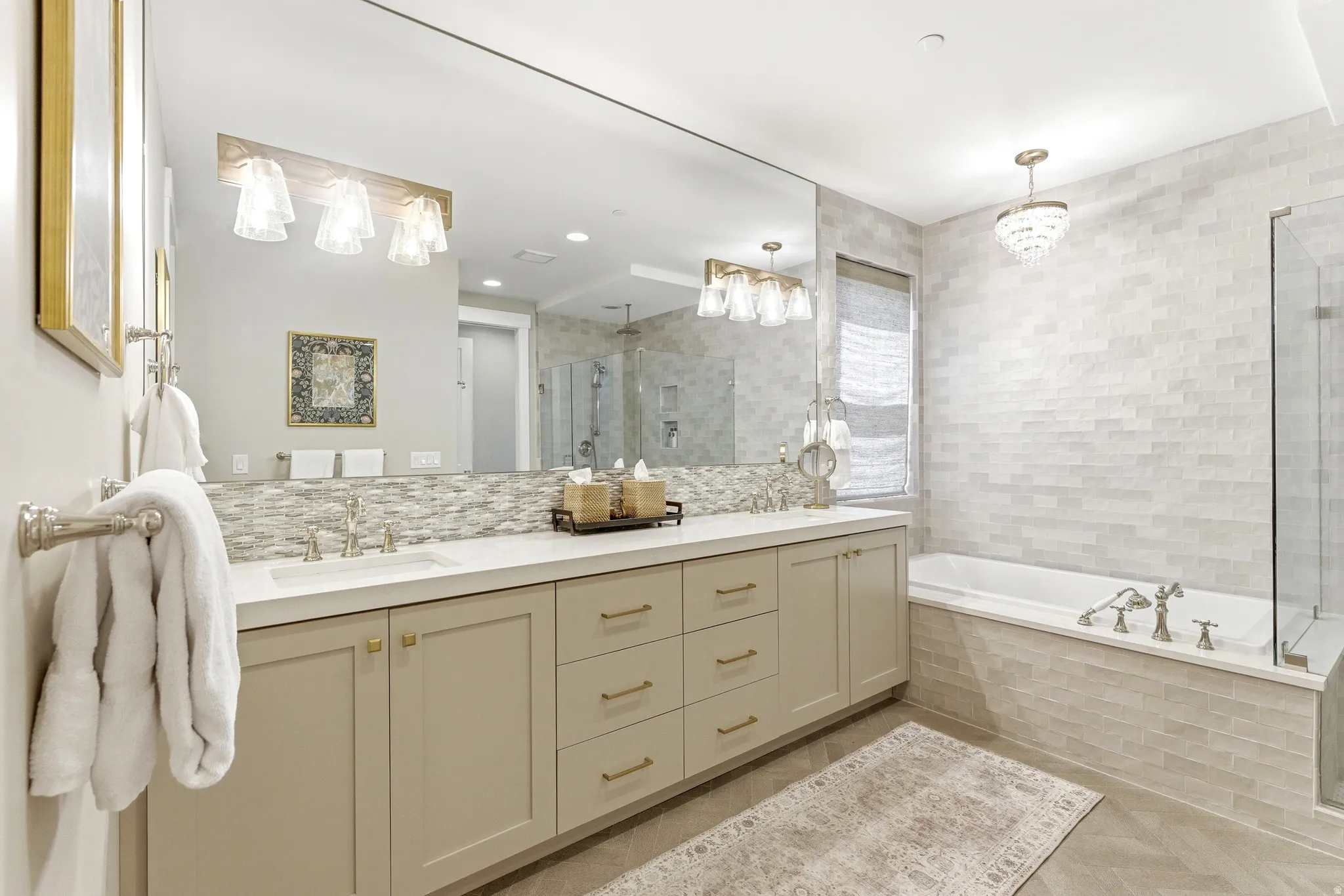 Bathroom featuring an extensive vanity with dual sinks, light-toned cabinetry with brass-finish hardware, a built-in soaking tub, a frameless glass shower enclosure, and light-toned tile work throughout