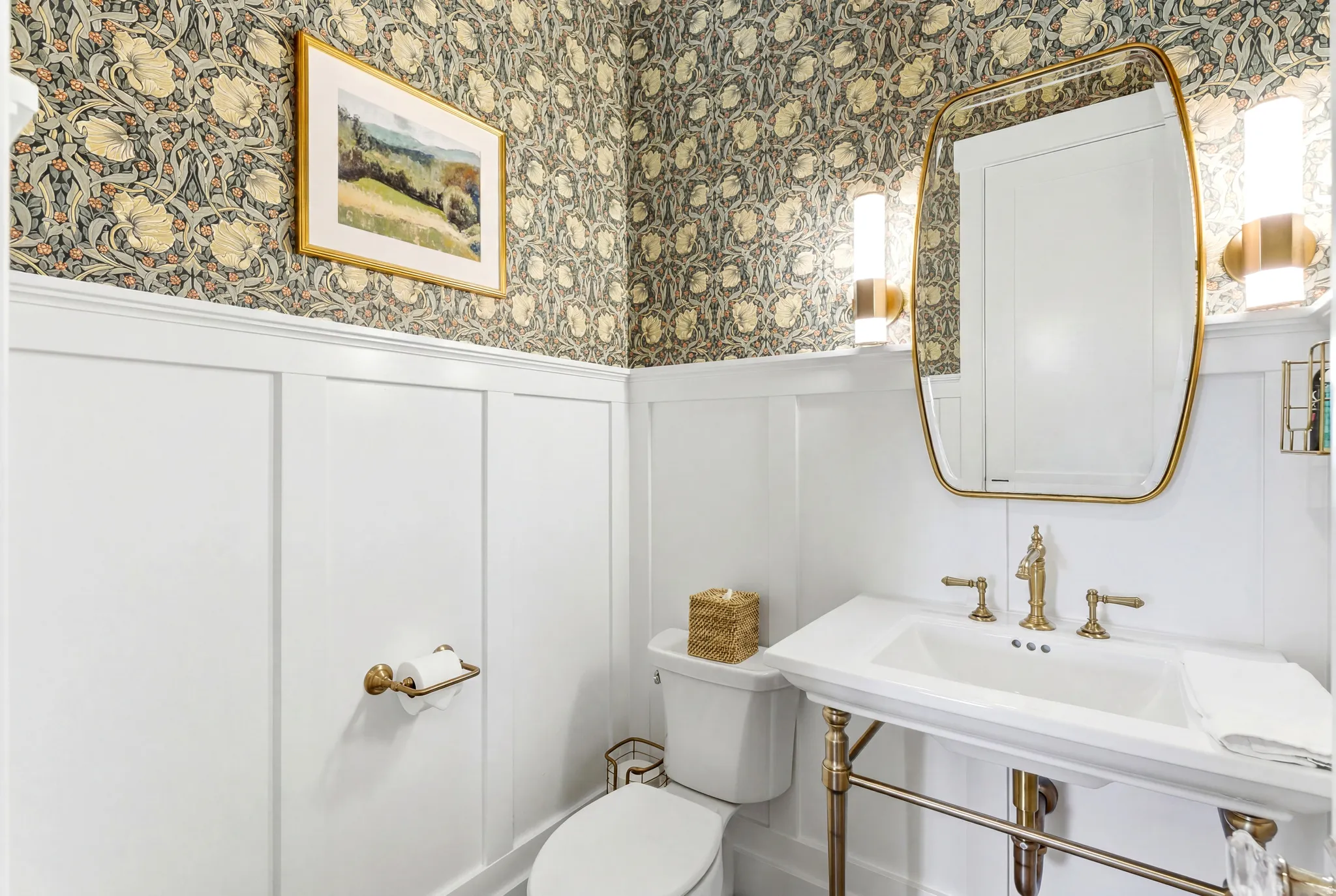 Powder room featuring board and batten wainscoting, botanical pattern wallpaper, and a pedestal sink with brass-finish fixtures