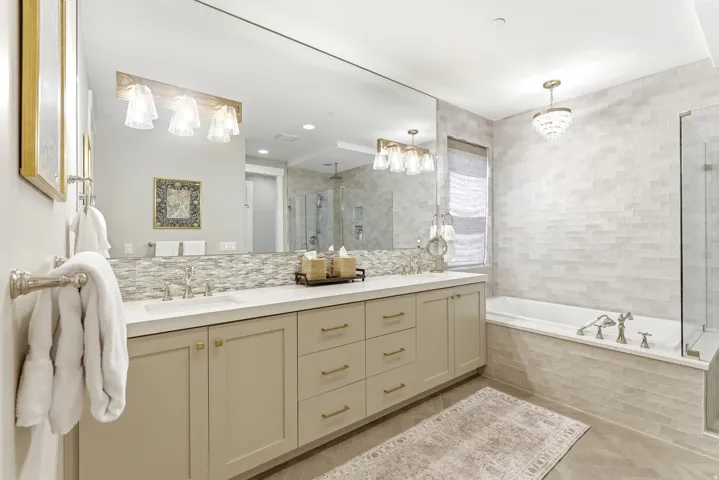 Bathroom featuring an extensive vanity with dual sinks, light-toned cabinetry with brass-finish hardware, a built-in soaking tub, a frameless glass shower enclosure, and light-toned tile work throughout