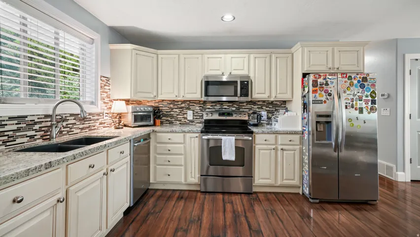 Kitchen featuring cream cabinetry, stainless steel appliances, granite countertops, mosaic tile backsplash, and walnut wood-finish flooring