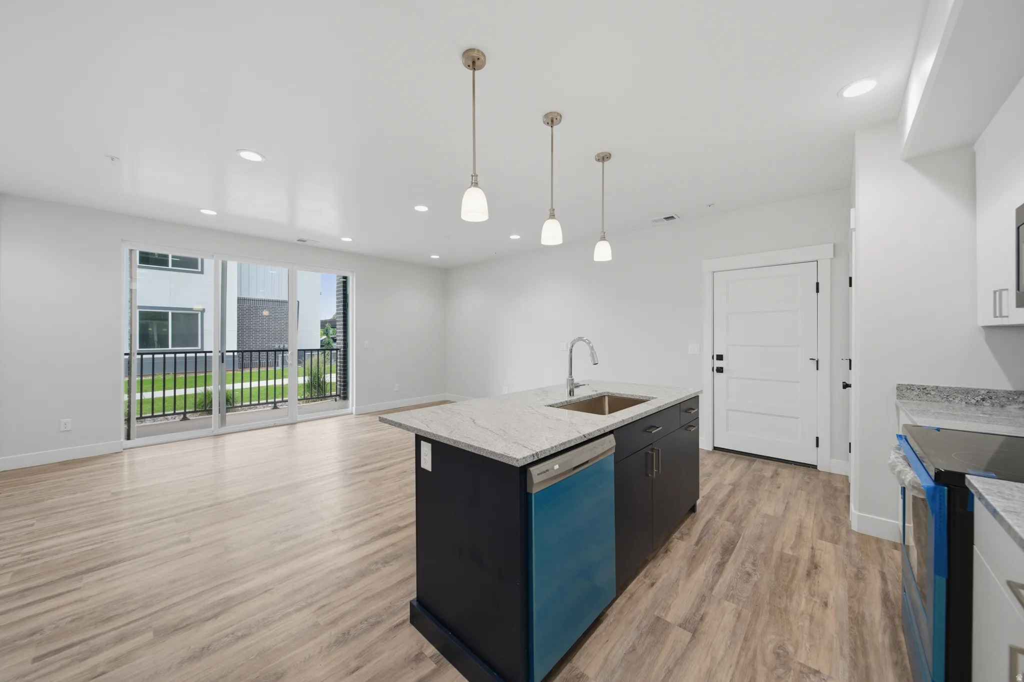 Open-concept living space featuring wood-finish flooring, a kitchen island with stone countertops and undermount sink, pendant lighting, recessed ceiling lights, and a sliding glass door leading to a balcony with black metal railings